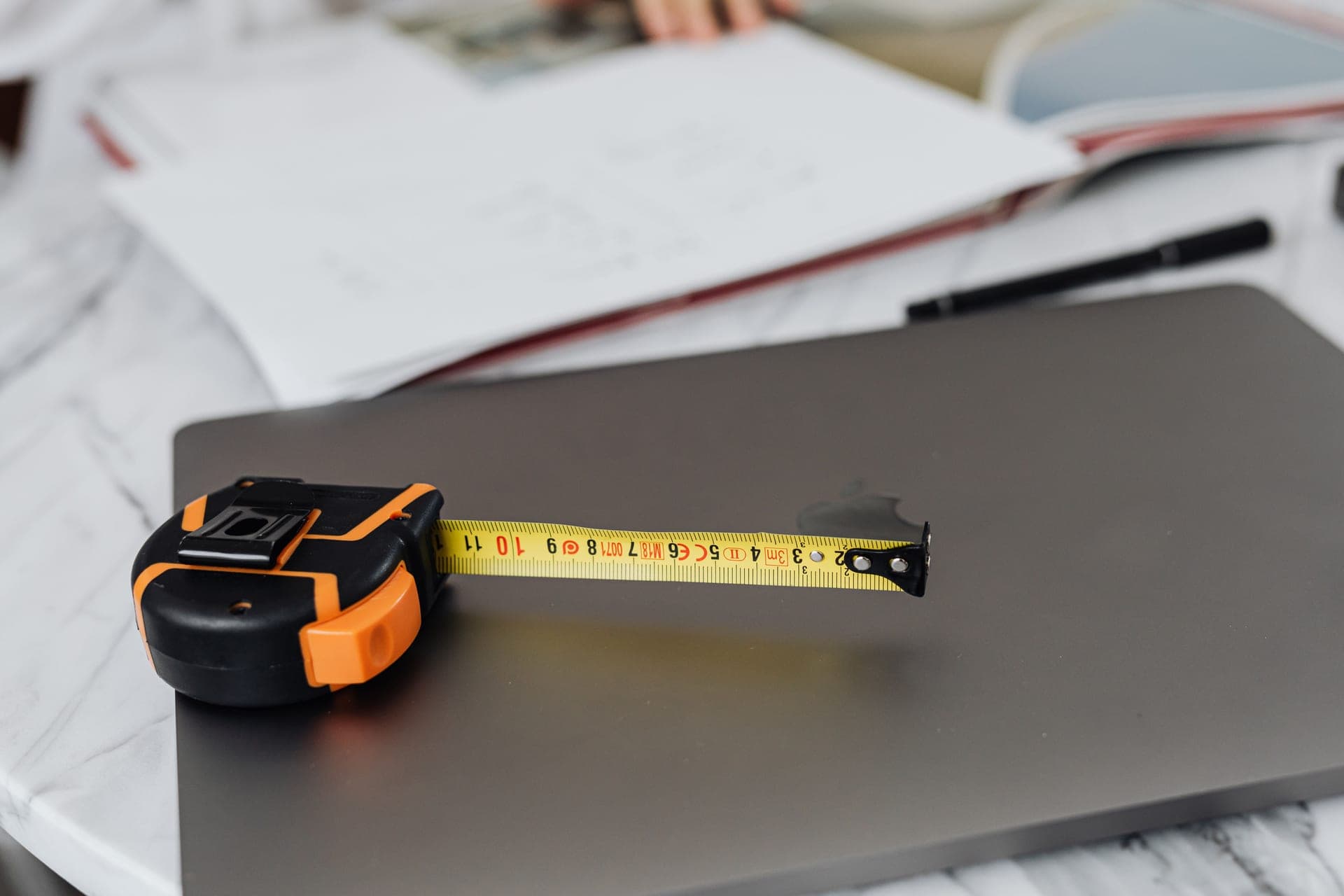 A notebook and tape measure on a table beside a window, with a room being measured in warm daylight